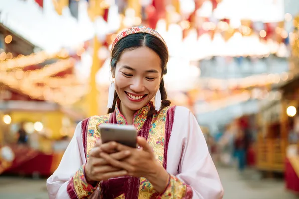 A cheerful woman in traditional clothing using her smartphone during a festive moment, showing how easily the CBAJI app fits into everyday life.
